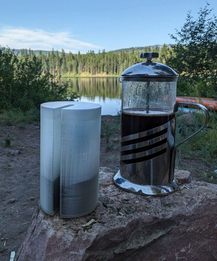 French press with Camel Joe canister on a red rock in front of a lake surrounded by forest